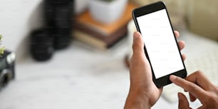 Imagen recortada de las manos de un hombre inteligente sosteniendo un teléfono inteligente negro recortado con una pantalla blanca en blanco sobre su escritorio de trabajo blanco como fondo.