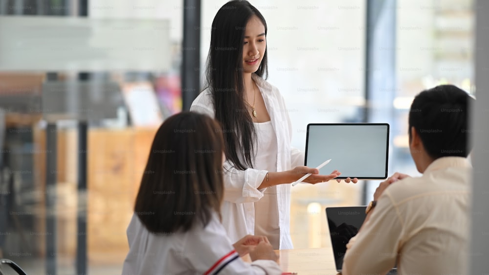 Photo of beautiful woman presenting her project with white blank screen computer tablet while standing in front her colleagues over modern meeting room as background.