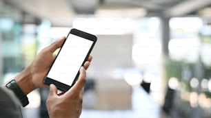 Cropped image of smart man's hands holding and using a cropped black smartphone with white blank screen over modern meeting room as background.