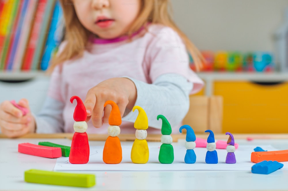 Bambina che modella gnomi colorati dall'argilla da gioco per bambini sul tavolo nella stanza dei bambini. Focus selettivo sugli gnomi. Attività domestiche per la famiglia.