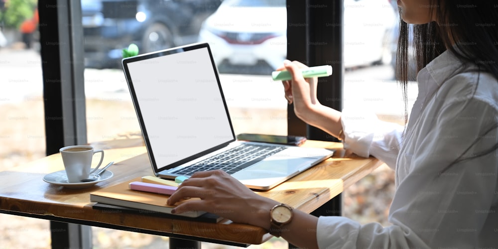 Cropped image of young secretary working with white blank screen laptop while sitting at wooden table over comfortable cafe glass wall as background. Working outside office concept.