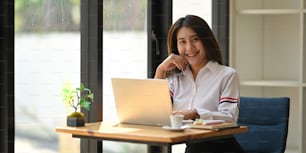 Attractive woman smiling and keep hand on chin while sitting in front her computer laptop at the wooden working desk over comfortable living room as background.