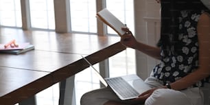 편안한 거실 위의 나무 작업 책상에 앉아 있는 동안 손에 책과 컴퓨터 노트북을 들고 있는 세련된 여성의 클로즈업 이미지.