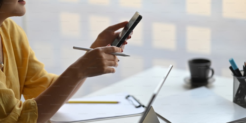 Foto de mulher elegante segurando um smartphone e caneta stylus nas mãos enquanto sentada na frente de seu tablet de computador com capa de teclado que coloca na mesa de trabalho branca sobre o escritório moderno como fundo.