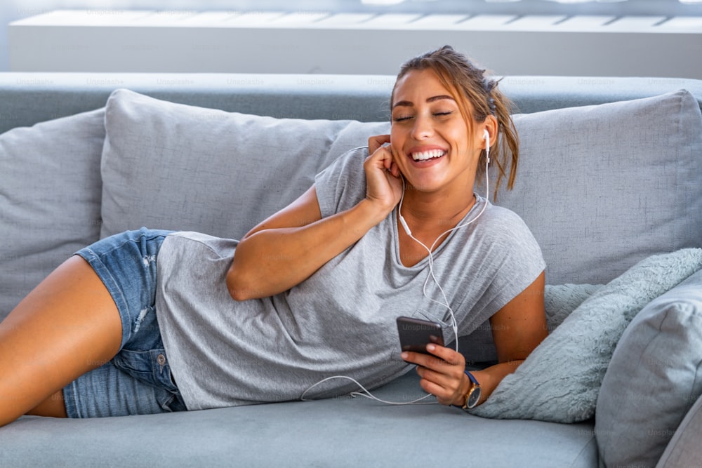 Relax in the rhythm of my music. Attractive woman with earphones and smartphone sitting on sofa at home. Beautiful girl in denim and headphones is listening to music Quarantine, Stay At Home concept
