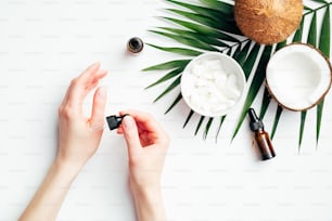 Female hands applying coconut oil for hand skin moisturizing. Flat lay composition with woman's hands, tropical palm leaf, coconuts, essential oils. Hand skincare, beauty treatment concept