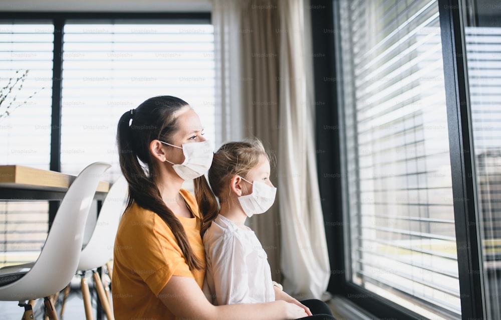 Mutter und kleines Kind mit Gesichtsmasken drinnen zu Hause, Blick nach draußen. Coronavirus und Quarantänekonzept.