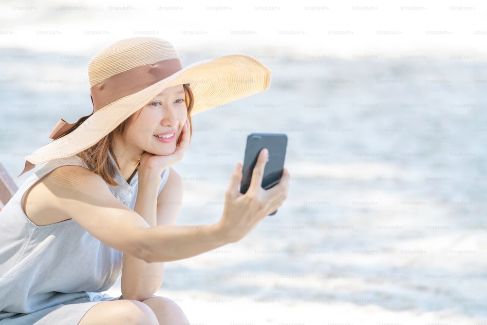 Joven hermosa mujer asiática sentada en silla de playa usando selfie o videollamada de teléfono inteligente