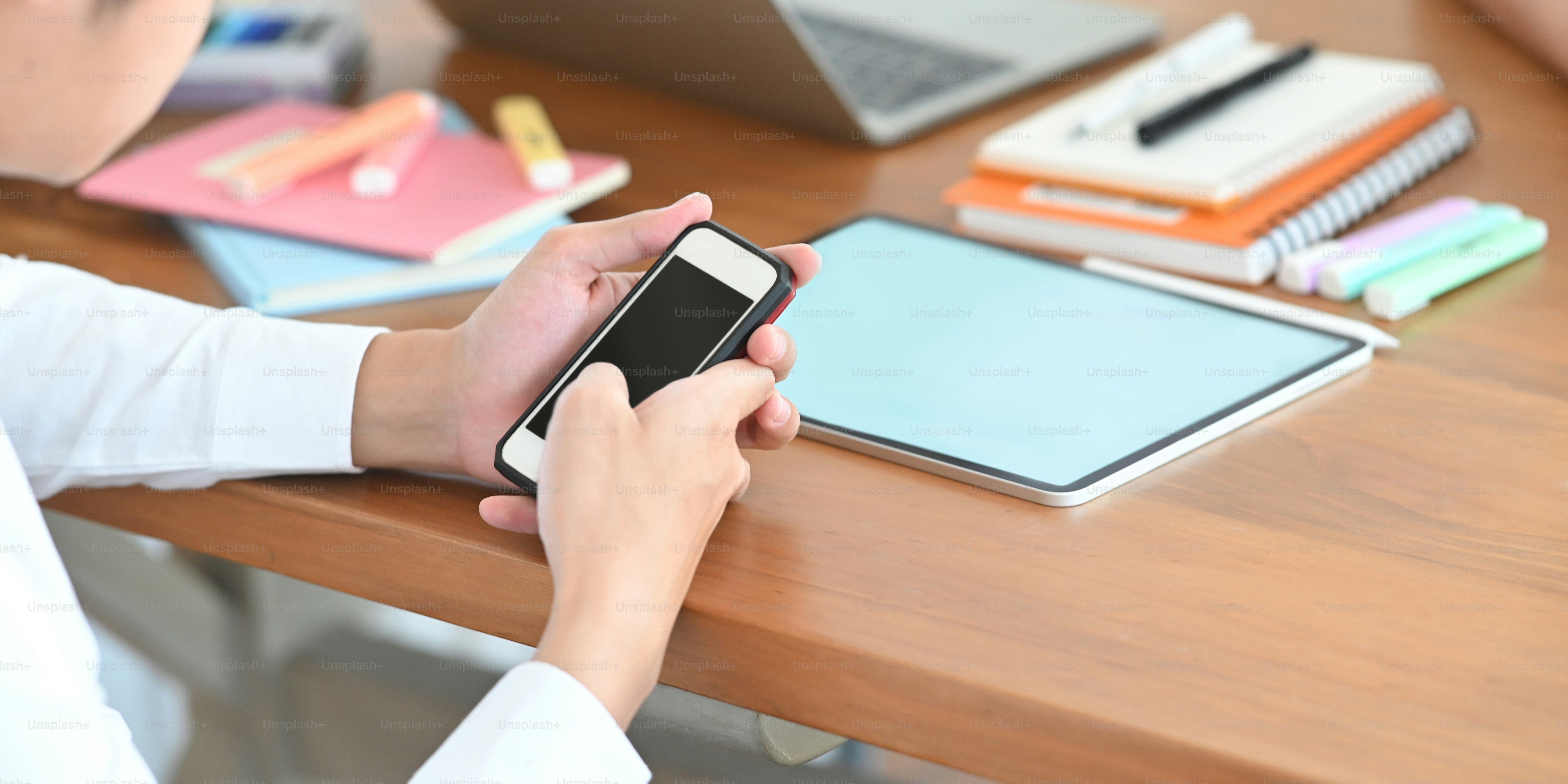 Immagine ravvicinata delle mani dell'uomo intelligente che tiene in mano uno smartphone con schermo vuoto nero mentre è seduto davanti al suo tablet con schermo bianco e penna stilo alla scrivania di lavoro in legno.