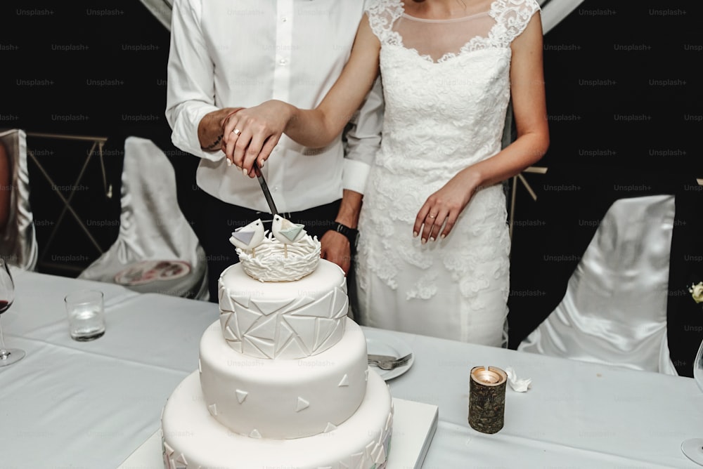 Novia hermosa con estilo y novio elegante cortando y degustando pastel de bodas blanco inusual