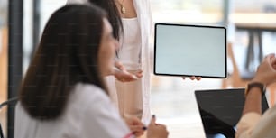 Mujer de primer plano que trabaja como marketing de pie y muestra una tableta de pantalla blanca en blanco en sus manos mientras se reúne con un colega en la mesa moderna sobre una sala de reuniones ordenada como fondo.