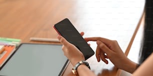 Close-up image of beautiful woman's hands holding a black blank screen smartphone in hands while sitting in front his computer tablet with white blank screen and stylus pen at the wooden working desk.