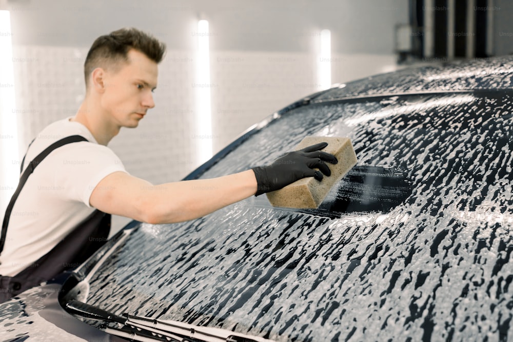 Junger männlicher Wascharbeiter reinigt die Windschutzscheibe des Autos mit Hilfe von speziellem Föm und gelbem Schwamm. Waschen des Autos von Hand, Schwamm und Schaumstoffblasen.