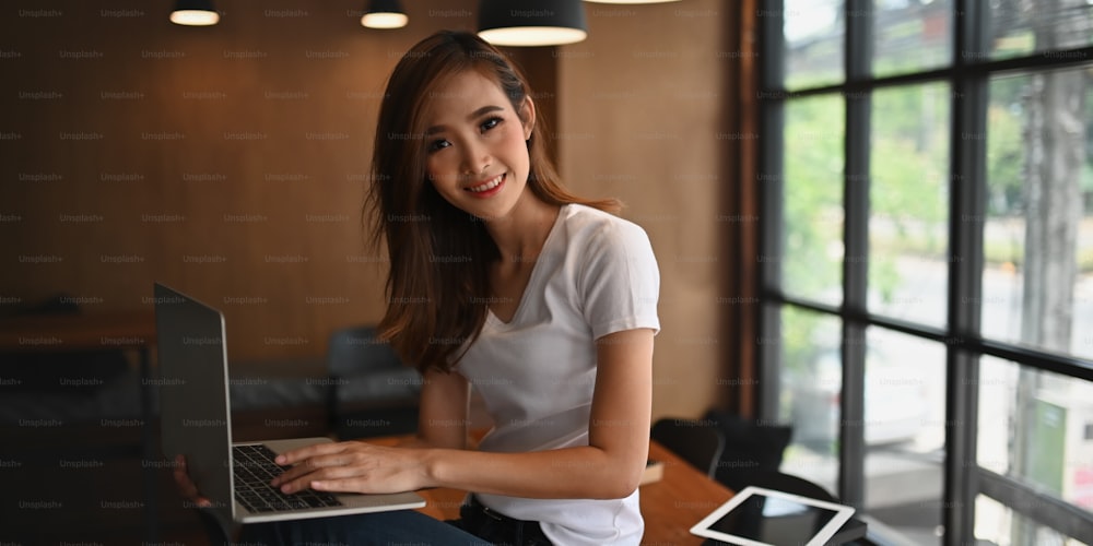 Photo of young entrepreneur woman smiling and sitting with her computer laptop that putting on her lap while relaxing in comfortable working room at home. Woman and relaxing time concept.