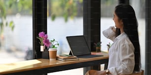 Geschäftsfrau streckt ihre Hand aus, während sie vor ihrem Computer-Tablet sitzt und sich entspannt, mit schwarzem leerem Bildschirm, der eine hölzerne Thekenbar über der Glaswand des Restaurants als Hintergrund anlegt.