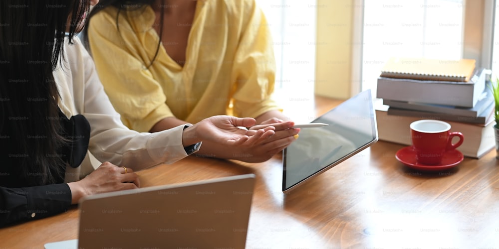 Cropped image of two beautiful women working together with computer laptop and tablet while sitting at the wooden working desk over comfortable living room as background.