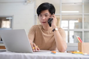 Junger männlicher Unternehmer, der mit einem Handy telefoniert und an einem Laptop arbeitet, während er an einem Tisch im Homeoffice sitzt.
