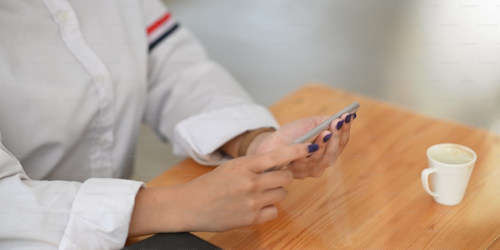 Cropped image of young woman's hands holding a smartphone while sitting at the wooden working desk over comfortable workplace as background. Woman resting time concept.