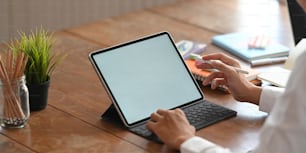 Cropped image of Graphic designer's hands holding a stylus pen and typing on computer tablet with keyboard case while sitting at the wooden working desk over orderly workplace as background.
