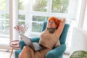 Tired elderly woman sitting in an armchair with laptop computer in her lap, stretching and yawning