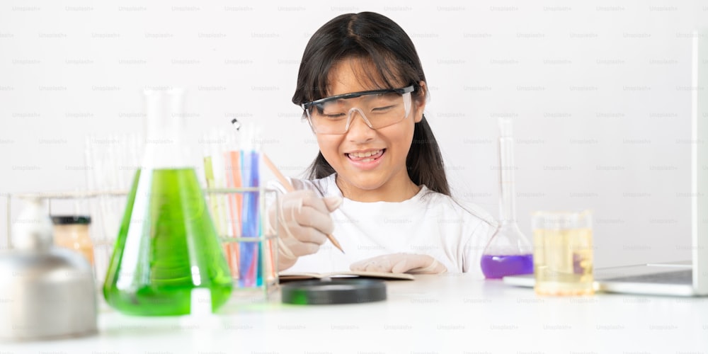Photo of young adorable school girl writing a scientific result while doing a scientific experiment and sitting at the modern white table with white laboratory wall as background.