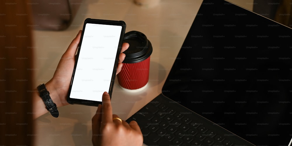Imagen recortada de manos sosteniendo un teléfono inteligente negro recortado con pantalla blanca en blanco sobre el escritorio de trabajo que está rodeado de equipos de oficina como fondo.
