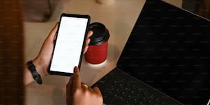 Cropped image of hands holding a cropped black smartphone with white blank screen over working desk that surrounded by office equipment as background.