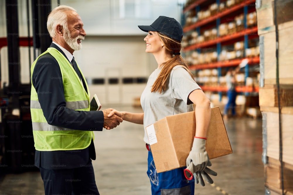 Happy female warehouse worker handshaking with company manager in industrial storage compartment.