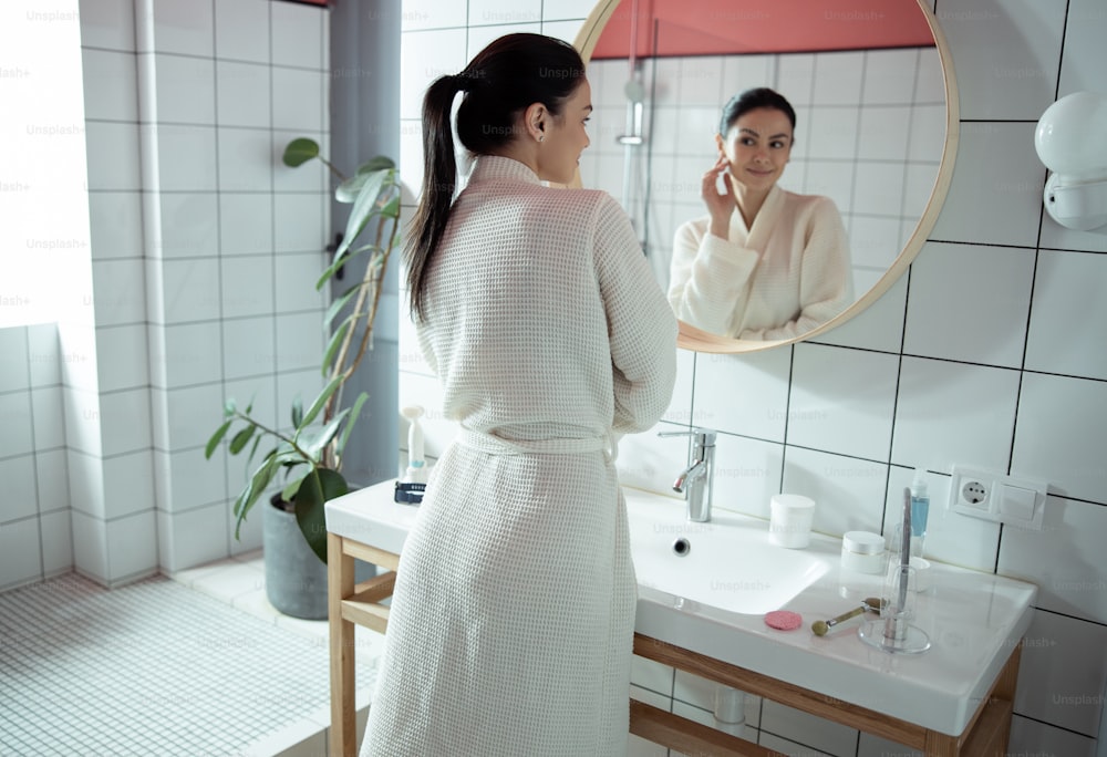 Merry young woman is wearing home bathrobe and standing in front of mirror while looking at herself