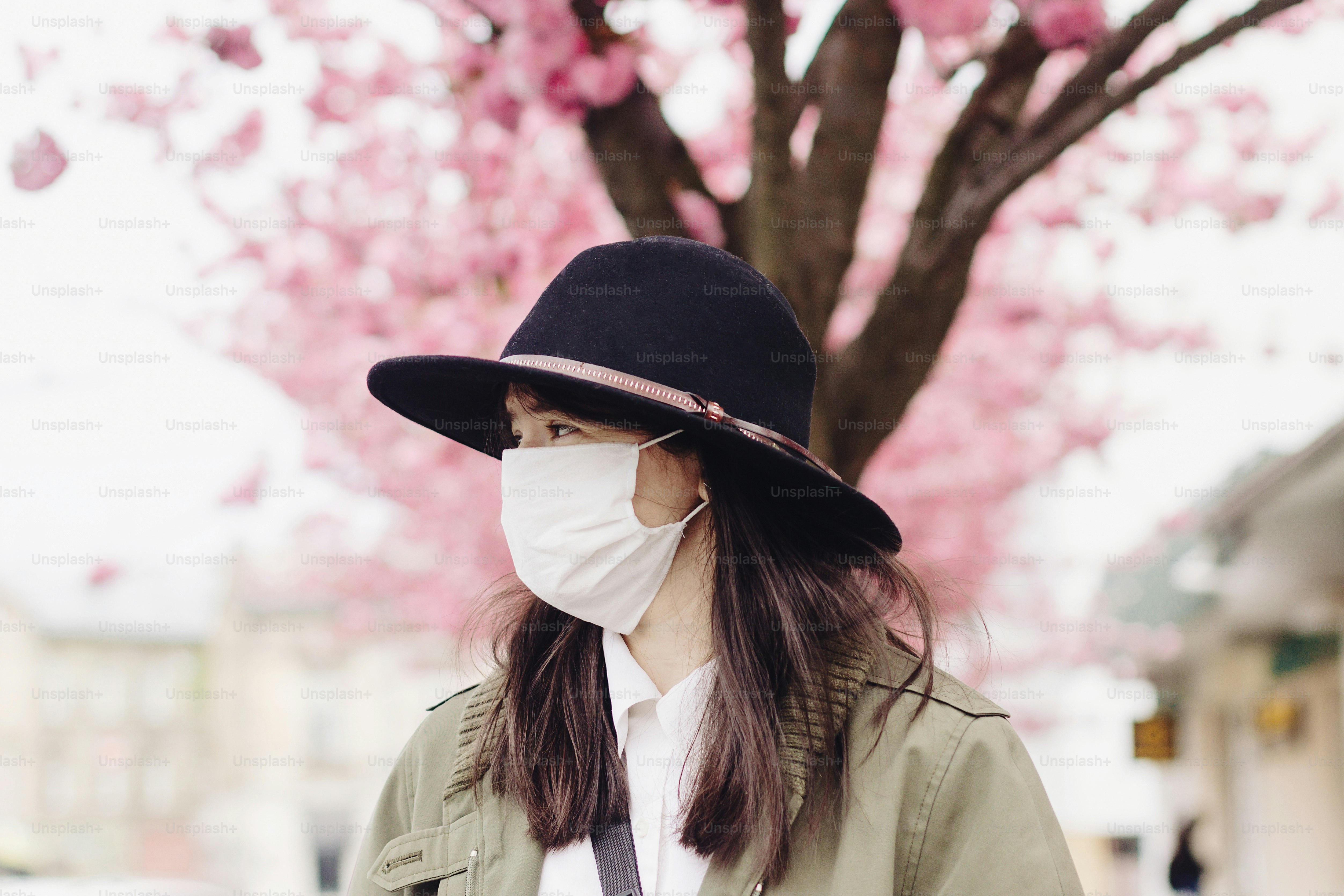 Sad hipster girl in face mask standing in city street on background of blooming pink trees during virus outbreak. Young woman wearing medical mask while walking outdoors