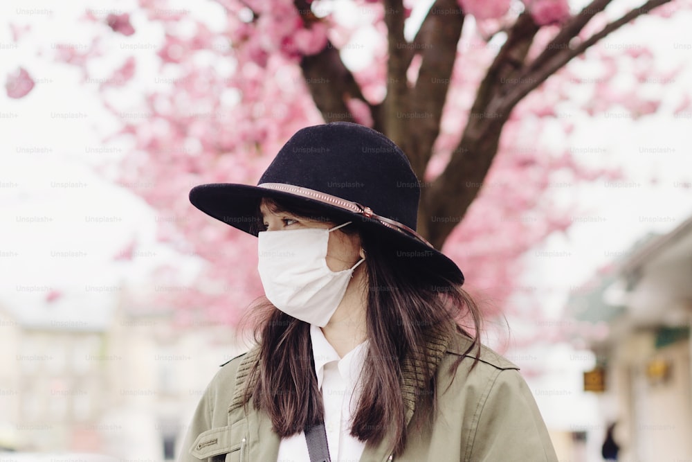 Sad hipster girl in face mask standing in city street on background of blooming pink trees during virus outbreak. Young woman wearing medical mask while walking outdoors