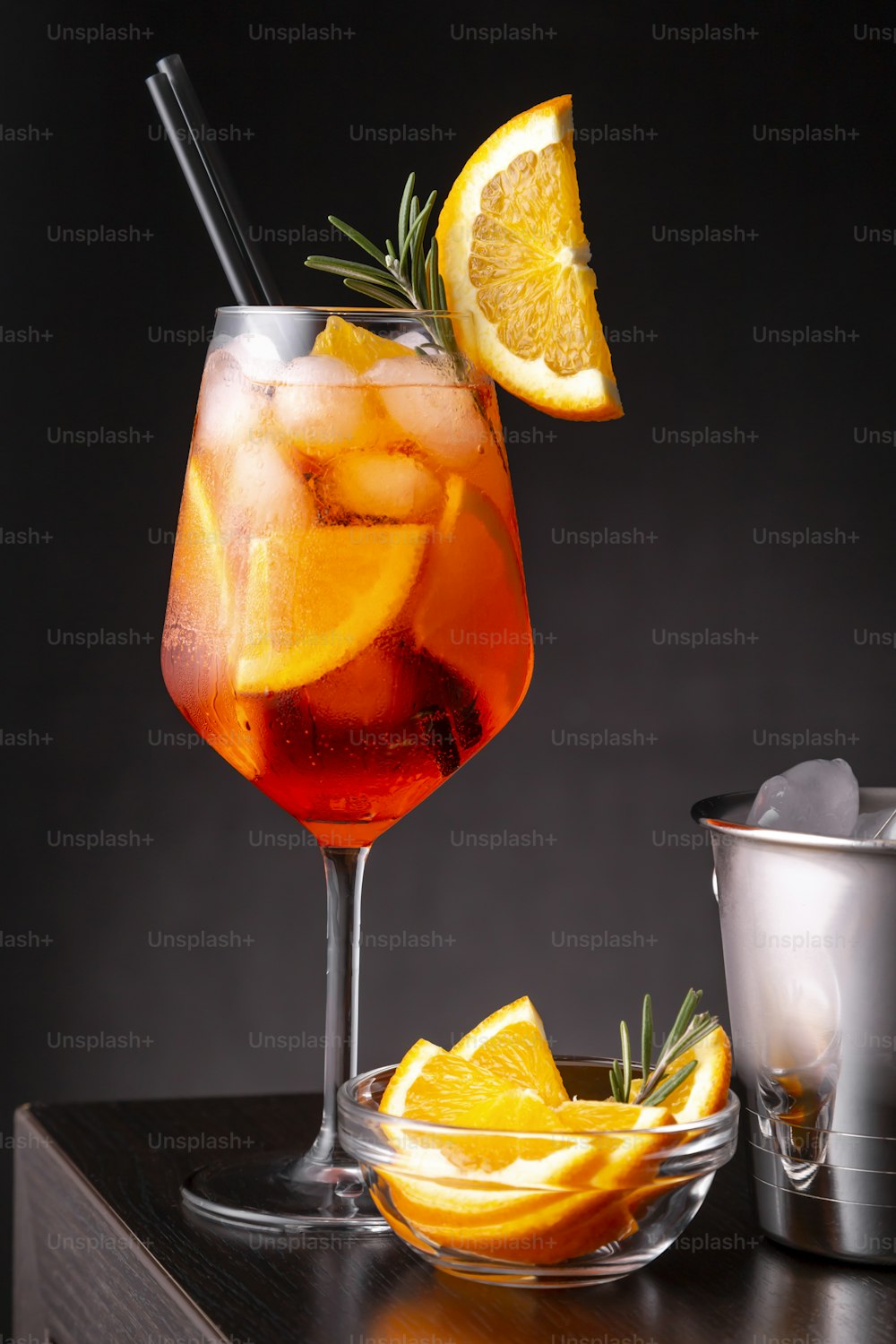 Spritz cocktail served in a wineglass with lots of ice, decorated with slice of orange and rosemary branch, placed on a bar counter