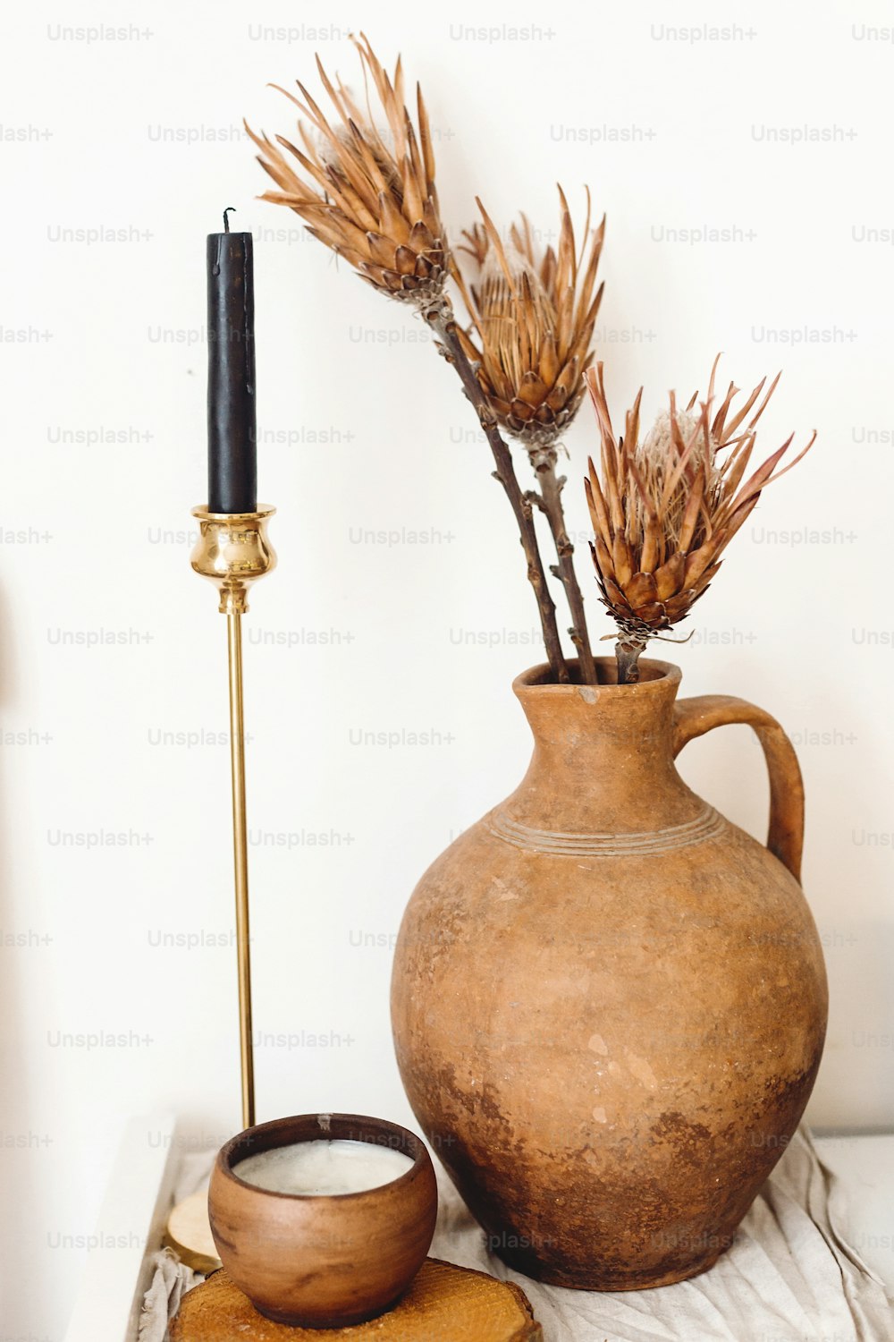 Dry protea flowers in old clay vase and candle on background of white wall in country home. Modern house decor. Stylish simple interior design elements. Rural house