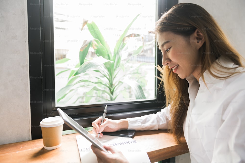 Young business woman in white dress sitting at table in cafe and writing in notebook. Asian woman using tablet and cup of coffee. Freelancer working in coffee shop. Student learning online.