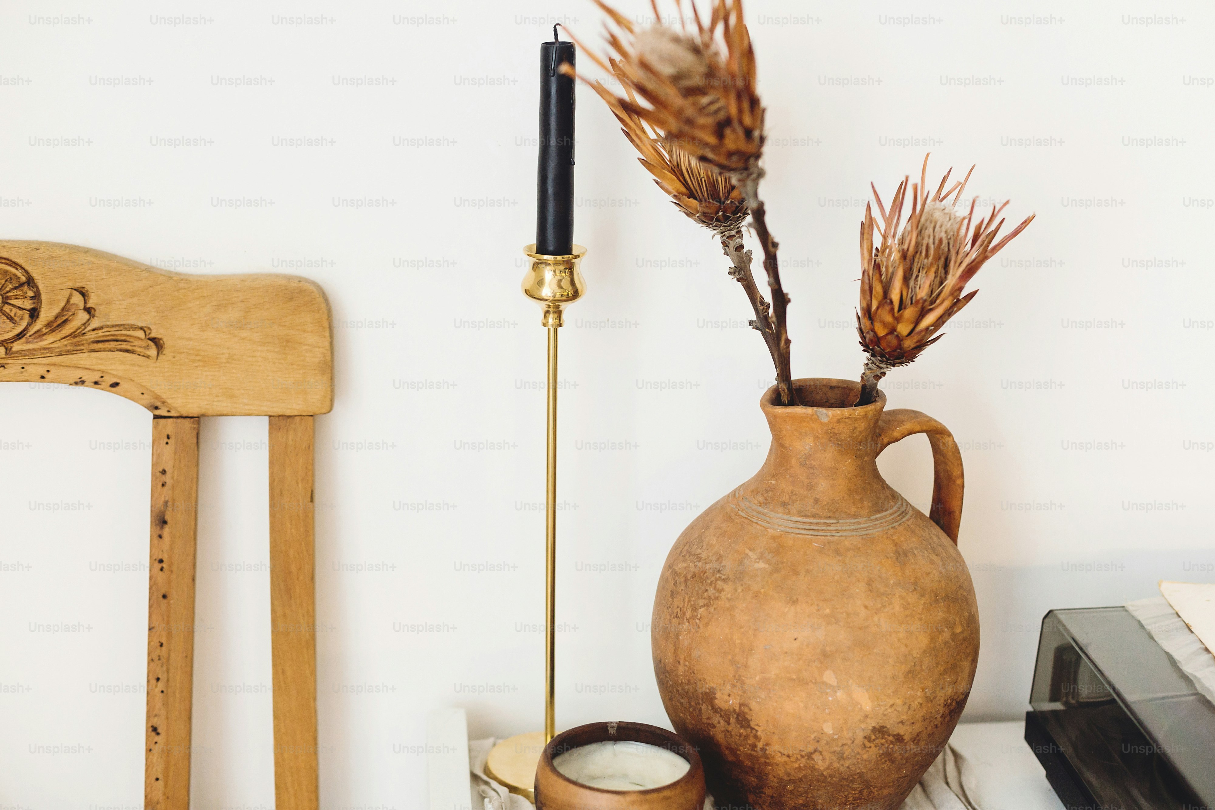 Bunga protea kering di vas tanah liat tua dan lilin dengan latar belakang dinding putih di rumah pedesaan. Dekorasi rumah modern. Elemen desain interior sederhana yang bergaya. Rumah pedesaan