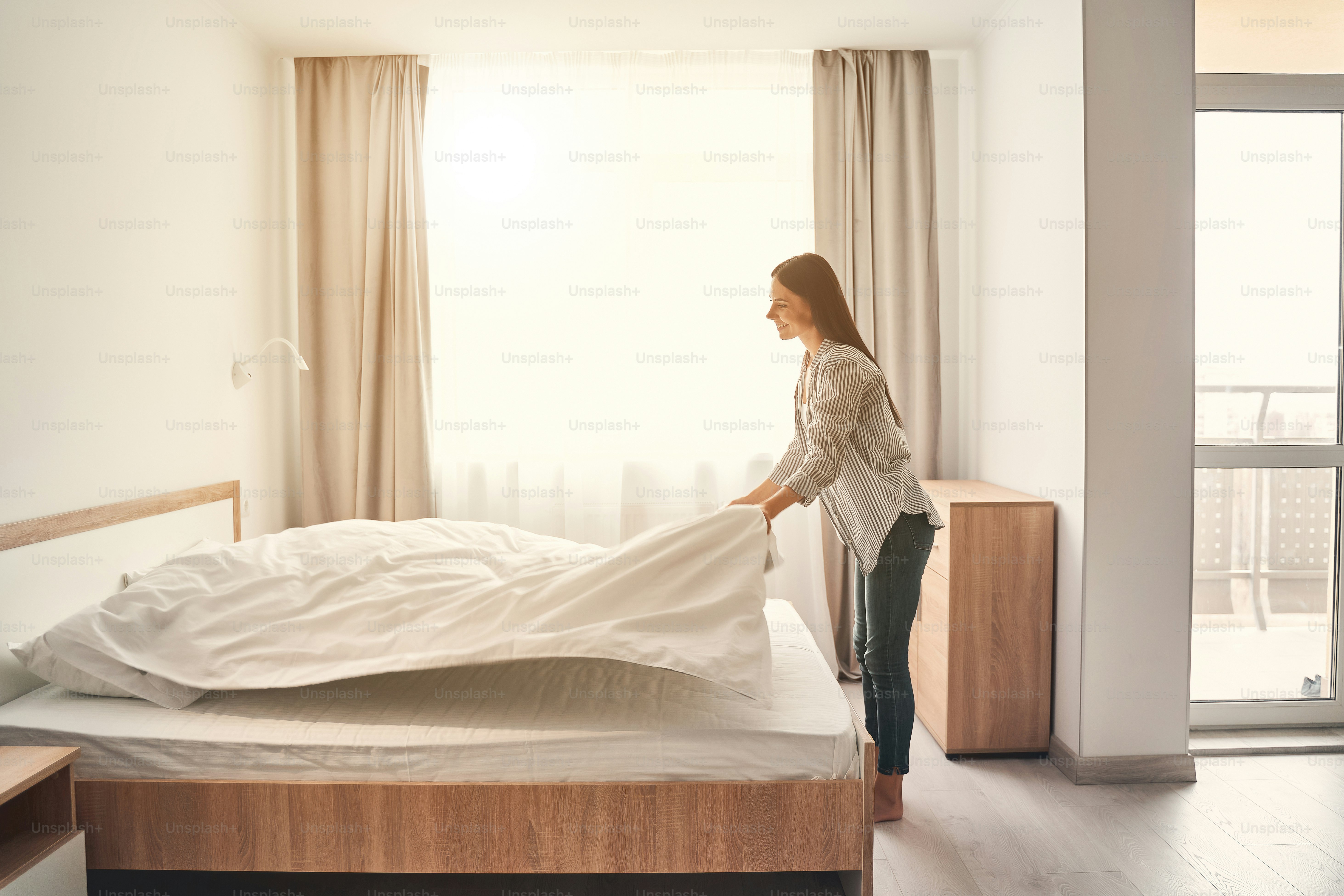 Fresh bedclothes. Pretty female person keeping smile on her face while making her bed