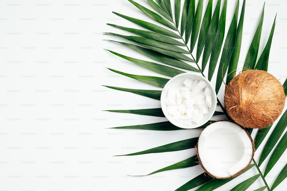 Coconut fruit with half and coconut flakes grated in bowl, tropical palm leaf on white background. Flat lay, top view. Summer background.