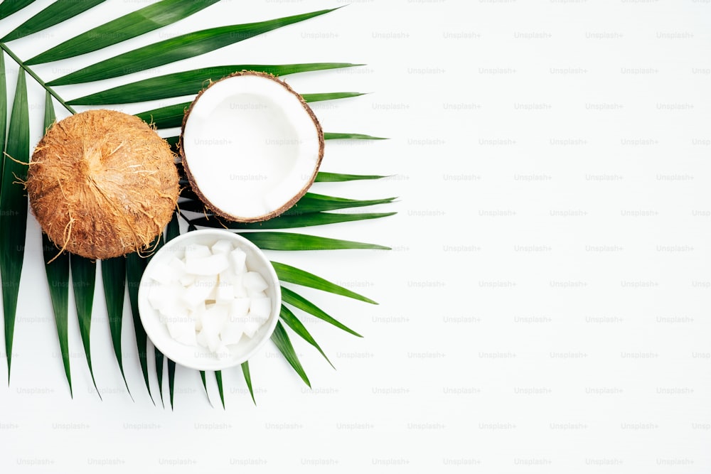 Coconut fruit with half and coconut flakes in bowl, tropical palm leaf on white background. Flat lay, top view. Summer background.