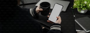Foto recortada de una mujer de negocios relajándose con café caliente y un teléfono inteligente de maqueta mientras está sentada en un lugar de trabajo moderno y oscuro