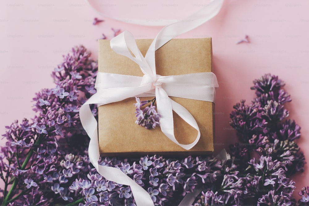 ピンクの背景にライラックの花とギフトボックス。スタイリッシュな花のグリーティングカード。紫色のライラックの花束、ピンクの紙にリボン付きのクラフトプレゼントボックス。母の日おめでとう