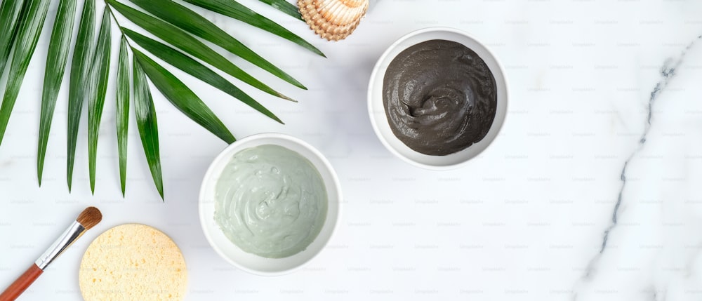 Green and black face clay masks in bowls with tropical palm leaf on marble background. Flat lay, top view. Facial skincare, SPA natural organic cosmetic concept