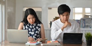 Young business couple working with computer laptop and tablet while sitting together at the long working desk over comfortable living room book shelf as background.