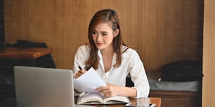 Foto de una mujer joven con estilo que usa una camisa blanca enfocada en su trabajo mientras está sentada frente a su computadora portátil en el escritorio de trabajo de madera sobre el lugar de trabajo contemporáneo como fondo.