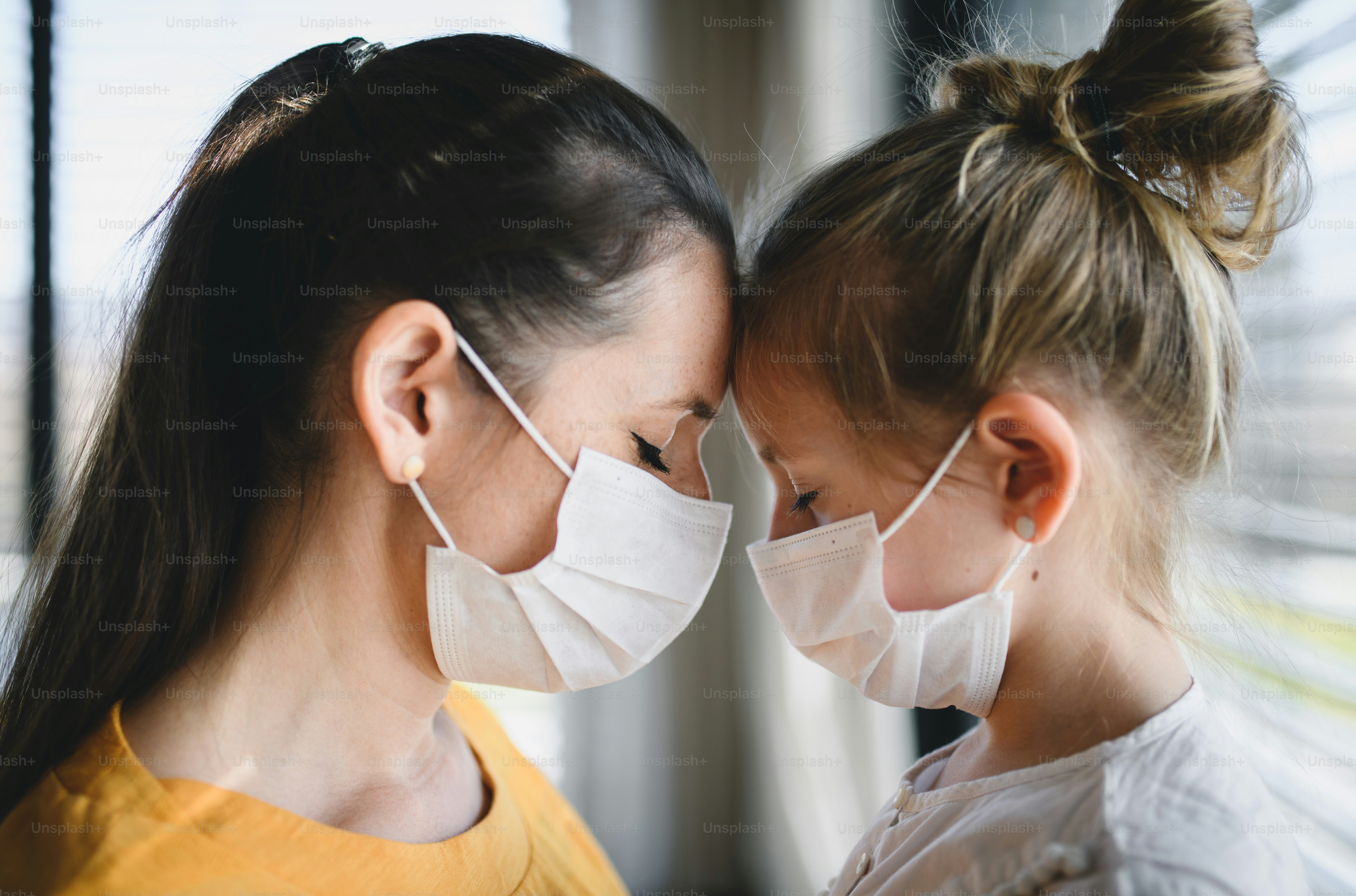 Mother and small child with face masks indoors at home, talking. Corona virus and quarantine concept.