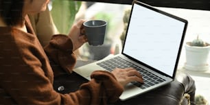 Cropped image of beautiful asian woman holding a coffee cup in hand while sitting in front her computer laptop with white blank screen that putting on her lap at the leather couch in living room.
