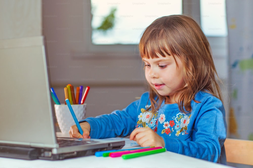 Bambina felice di quattro anni che fa i compiti usando il quaderno per l'apprendimento a distanza seduta al tavolo nella stanza dei bambini. Concetto di apprendimento online a distanza.