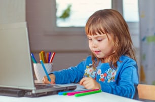 Heureuse petite fille de quatre ans faisant ses devoirs à l’aide du cahier pour l’apprentissage à distance, assise à la table dans la chambre des enfants. Concept d’apprentissage en ligne à distance.