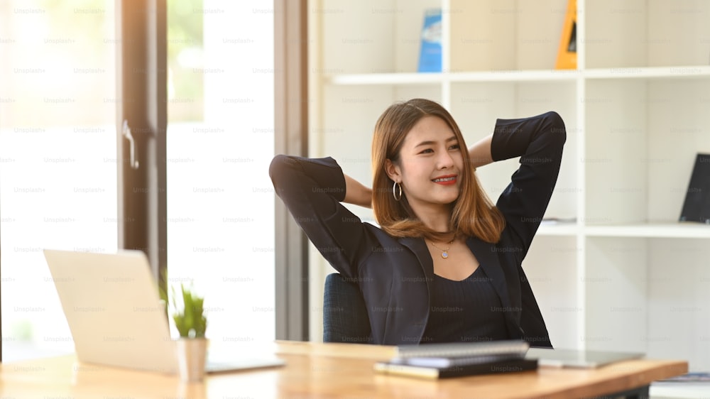 Gorgeous office woman relaxing by stretching her body while sitting in front her computer laptop at the wooden working desk over comfortable living room as background.