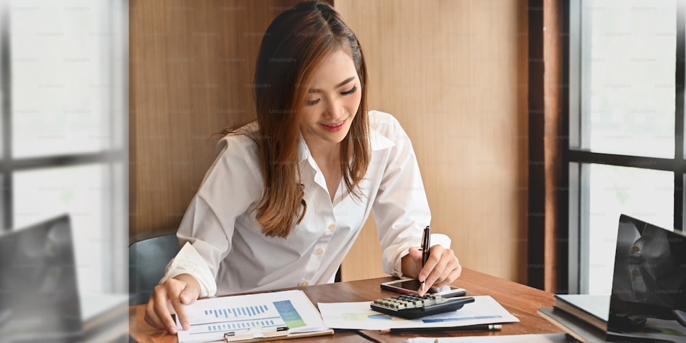 Photo of young stylish woman wearing on white shirt focused on her work while sitting in front her computer laptop at the wooden working desk over contemporary workplace as background.