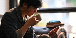 Cropped image of smart man drinking a hot coffee and holding/using a white blank screen smartphone while sitting and relaxing at the black leather sofa over comfortable sitting room as background.